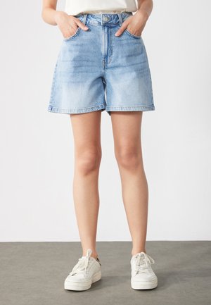 Persoană purtând pantaloni scurți din denim albastru deschis și adidași albi, stând cu mâinile în buzunare pe un fundal simplu.