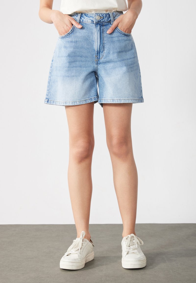 Asmuo dėvintis šviesiai melsvus džinsinius šortus ir baltus sportinius batelius, stovintis su rankomis kišenėse prieš paprastą foną.