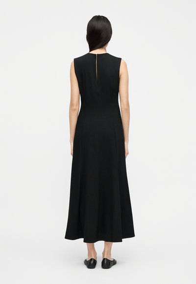 Naine seisab seljaga vaataja poole, kandes varrukateta musta pahkluudeni kleiti, millel on seljal lõhik, ning musta madala kontsaga jalanõusid.