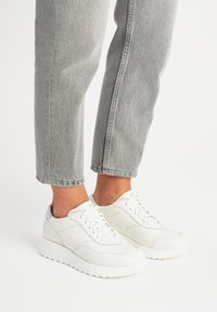 Białe skórzane trampki z teksturowanymi akcentami, zaokrąglonym palcem i sznurowaniem, w zestawieniu z szarymi, krótkimi dżinsami.