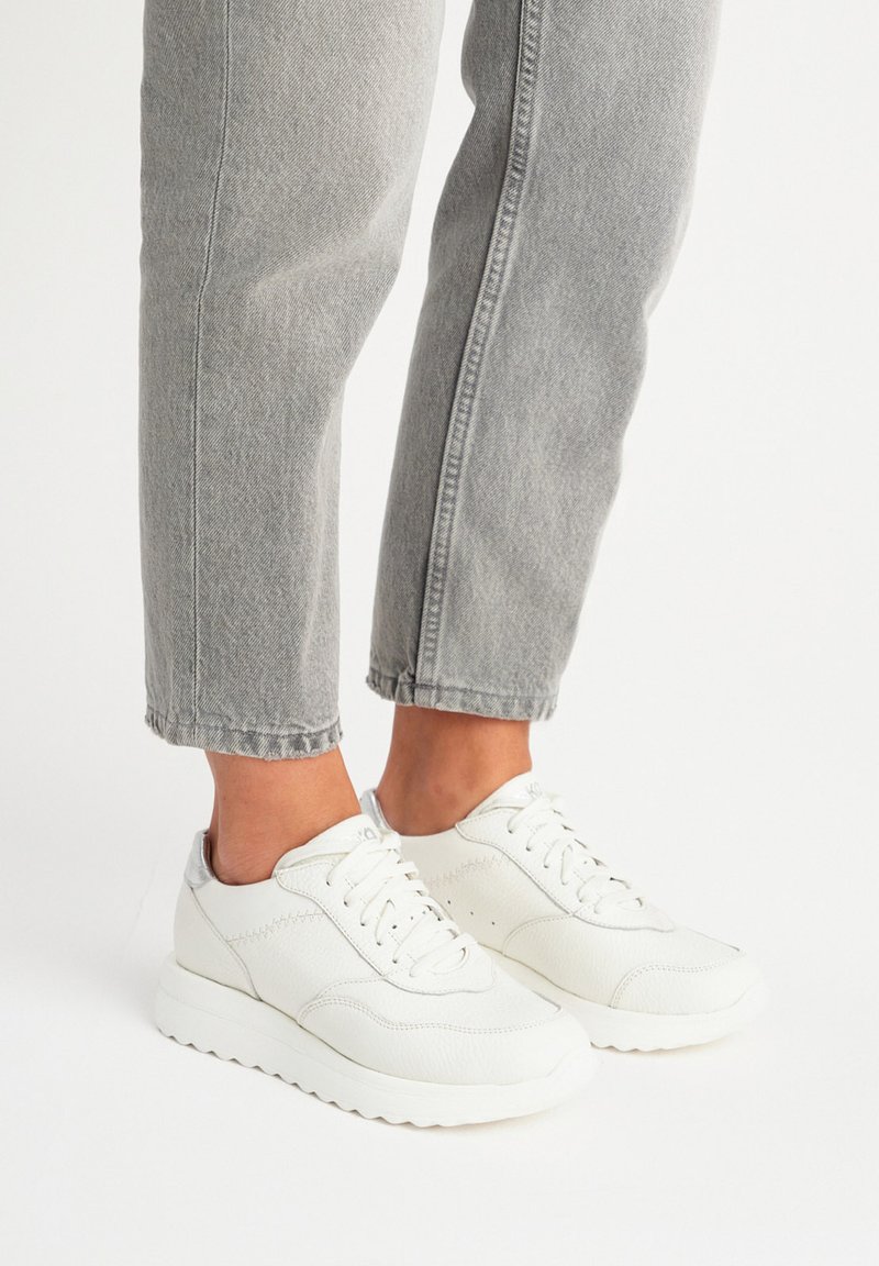 Białe skórzane trampki z teksturowanymi akcentami, zaokrąglonym palcem i sznurowaniem, w zestawieniu z szarymi, krótkimi dżinsami.