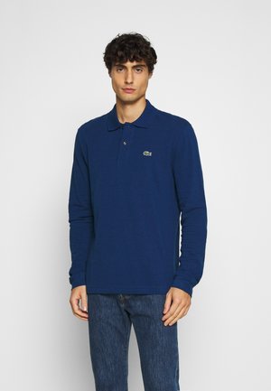 Camisa polo de manga larga en color navy con cuello, dos botones y un pequeño logo verde bordado en el pecho. Se lleva con unos vaqueros de mezclilla azules.