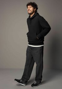 Schwarzer Kapuzenpullover mit Fronttasche, über einem weißen T-Shirt getragen und kombiniert mit dunkelgrauen Jeans und schwarzen Sneakers. Lockige Frisur.