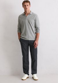 Graues langärmliges Polo-Shirt mit Kragen, kombiniert mit dunkelbraunen maßgeschneiderten Hosen und weißen Turnschuhen, vor einem hellen Hintergrund.