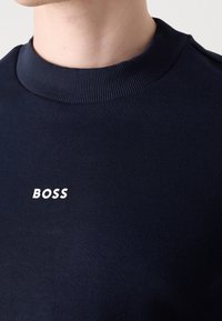 Gros plan d'une personne portant un pull bleu marine foncé à col rond côtelé avec le texte blanc « BOSS » sur la poitrine.