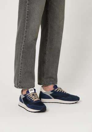 Sneaker navy con accenti grigi e crema, materiali in camoscio e mesh, punta rotonda e lacci beige, abbinati a pantaloni in denim grigi.