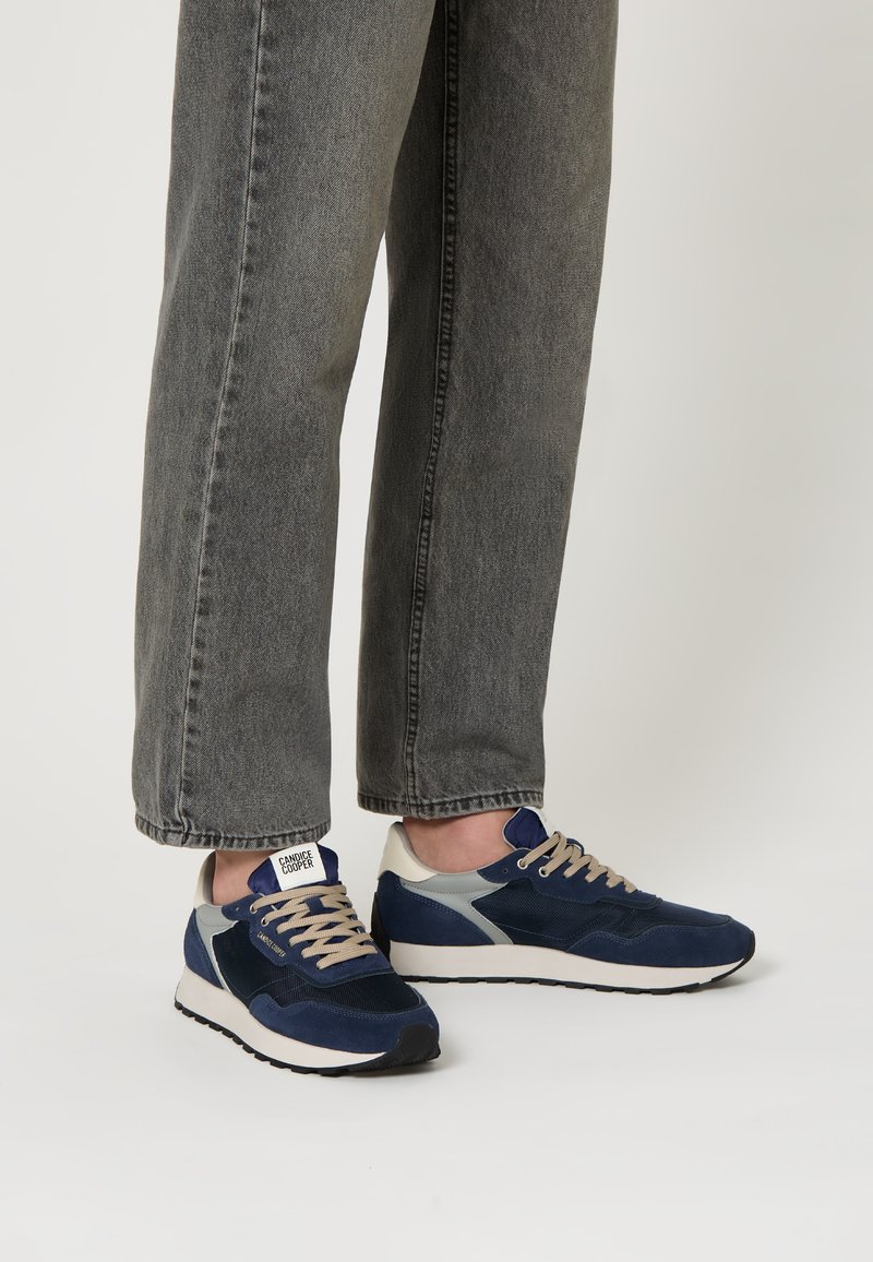 Sneaker navy con accenti grigi e crema, materiali in camoscio e mesh, punta rotonda e lacci beige, abbinati a pantaloni in denim grigi.