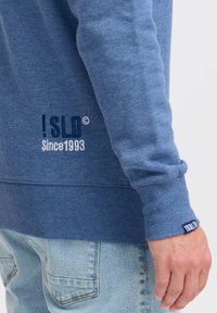Blue sweatshirt in soft fabric, featuring ribbed cuffs, embroidered logo in dark blue and white, and a relaxed fit with a textured hem.