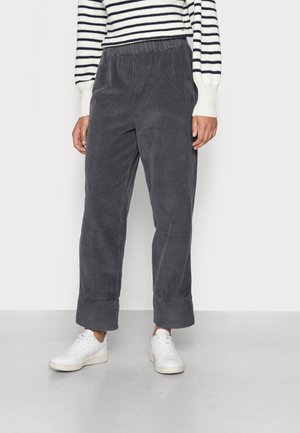 Personne portant un pantalon large en velours côtelé gris foncé avec des revers roulés, des baskets blanches et un pull blanc avec des rayures horizontales noires.