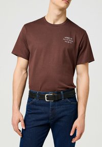 Camiseta de algodón marrón de manga corta, con texto bordado en blanco. Combinada con jeans de color azul oscuro y un cinturón de cuero negro con hebilla plateada.