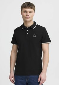 Svart polo skjorte med hvit kant rundt kragen og ermene, med tre knapper og en liten logo på venstre bryst.