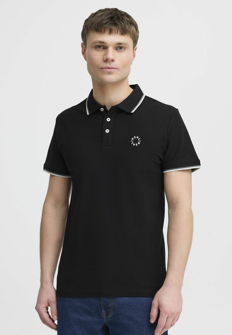 Svart polo skjorte med hvit kant rundt kragen og ermene, med tre knapper og en liten logo på venstre bryst.