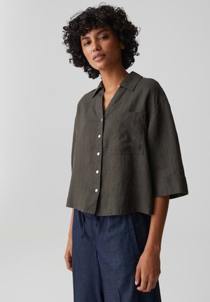 Frau mit lockigem Haar trägt ein lockeres, dunkelolivfarbenes Hemd mit Dreiviertelärmeln und blaue weit geschnittene Hosen, steht vor einem schlichten Hintergrund.