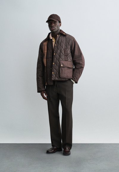 Άνδρας που φοράει καφέ μπουφάν με καπιτονέ ραφές, καπέλο σε ίδιο καφέ χρώμα, σκούρο καφέ παντελόνι και καφέ δερμάτινα παπούτσια, στέκεται μπροστά σε ένα απλό ανοιχτό φόντο.