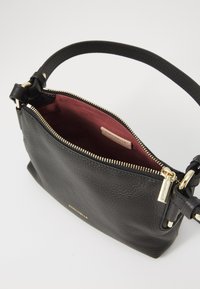 Borsa a tracolla in pelle nera con superficie texturizzata, zip dorata e fodera bordeaux. Include una tracolla regolabile e un piccolo etichetta con logo.