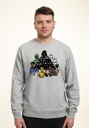 Mand iført en grå sweatshirt med Star Wars-figurer inklusive Darth Vader, stormtroopers, Chewbacca og droider, stående mod en ensfarvet baggrund.