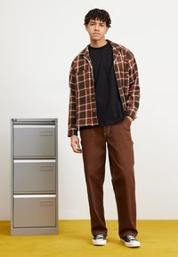 Brunternetternet skjortejakke over en sort t-shirt, parret med brune bukser og sorte sneakers, står ved siden af et gråt arkivskab på gule gulve.