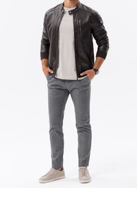 Braune Leder-Fliegerjacke, hellgraues T-Shirt, graue maßgeschneiderte Hose und beige Sneakers. Das Model posiert mit den Händen in den Taschen.