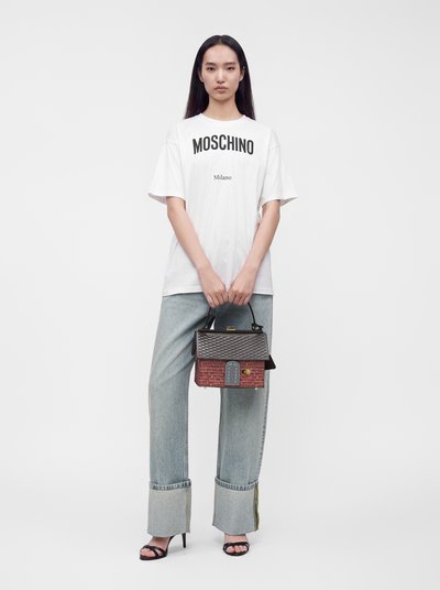 Femme portant un t-shirt blanc Moschino, jean bleu clair à larges revers, sandales noires, tenant un sac à main en forme de maison en brique avec toit gris et fenêtre.