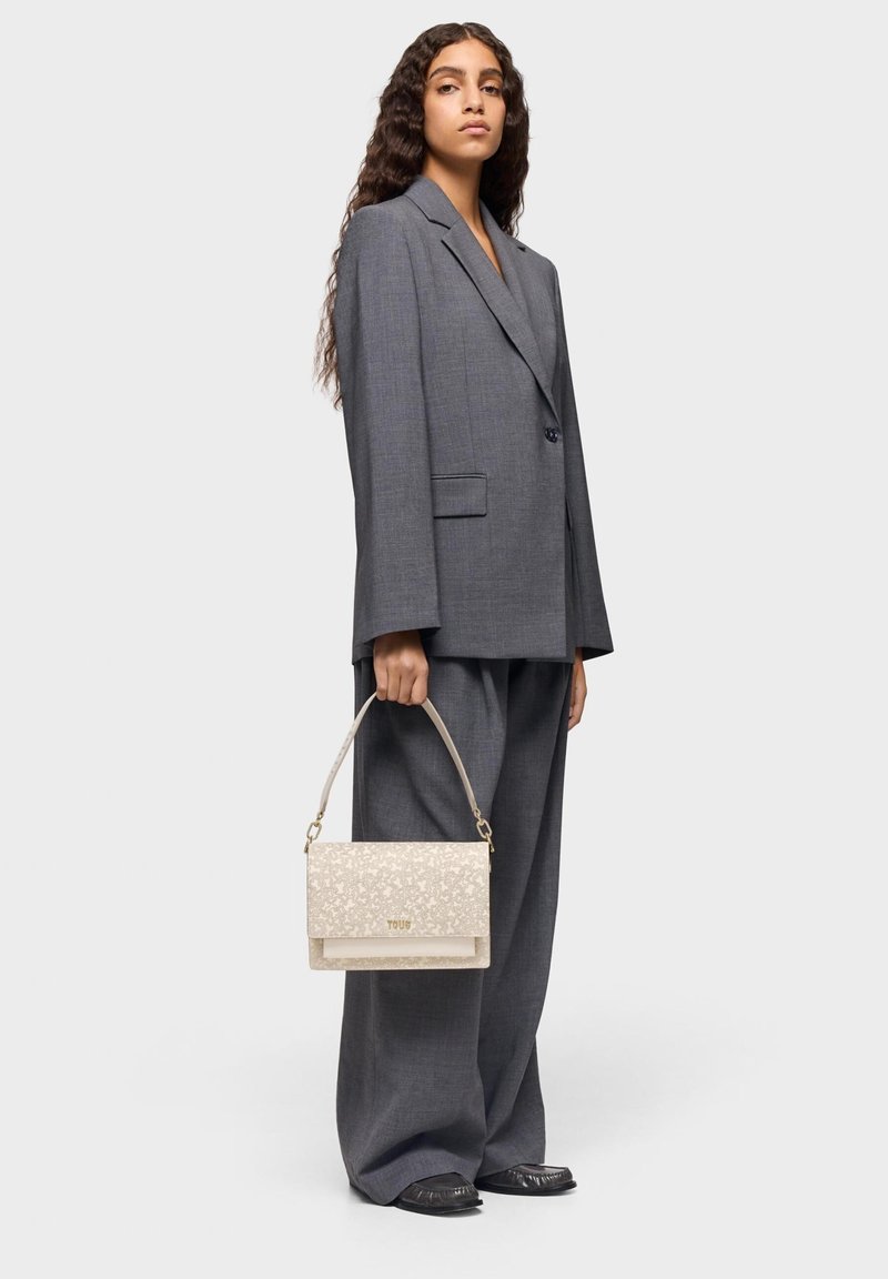 Gray oversized blazer and pants worn by a model, holding a beige textured handbag with a floral pattern and gold accents.