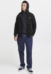 Schwarze Fleecejacke mit Reißverschluss und Brusttasche über einem marineblauen Hoodie, kombiniert mit marineblauen Hosen und schwarzen Sneakers.