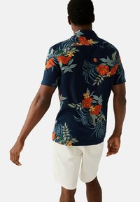 Camicia a maniche corte di colore blu scuro con motivo floreale rosso e giallo, con foglie verdi. Abbinata a dei pantaloni corti di colore chiaro.