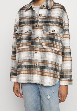 Person iført en brun, sort og hvid ternet skjorte med knapper og brystlommer, kombineret med lyseblå slidte jeans.