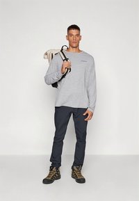 Hallitriibuline hall pluus ees oleva logoga, tumesiniste pükstega ja vastupidavate mustade ja kollaste matkasaabastega, kandes beež seljakotti.