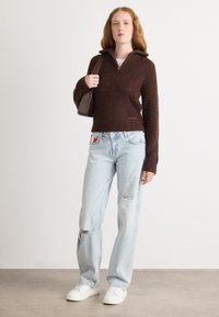 Bruine geribde gebreide trui met een ritskraag, lichtblauwe versleten jeans en witte sneakers. Model houdt een donkere handtas vast.