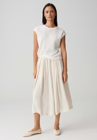 Witte, gestructureerde top met korte mouwen gecombineerd met een vloeiende, crèmekleurige midirok. Zacht materiaal en een eenvoudig ontwerp. Neutrale ballet flats maken de look compleet.