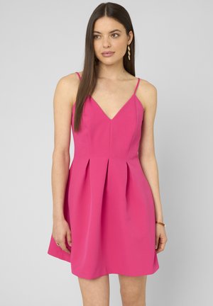 Femme aux longs cheveux bruns portant une robe plissée rose vif sans manches à décolleté en V, debout devant un fond gris clair uni.