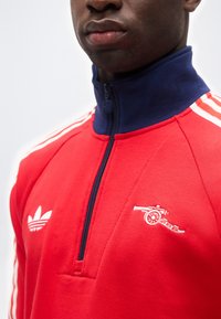 Κοντινό πλάνο ατόμου που φοράει κόκκινο ανδρικό φερμουάρ Adidas με ναυτικό γιακά και λευκά κεντημένα λογότυπα Adidas και κανόνι στο στήθος.