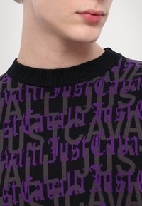 Close-up of a person’s lower face and neck wearing a black sweater with purple and gray "Just Cavalli" text pattern.