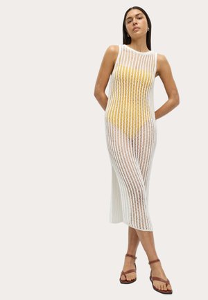 Abito bianco senza maniche in maglia con trama verticale aperta, di lunghezza fino al polpaccio, indossato sopra un costume da bagno giallo, completato da sandali marroni.