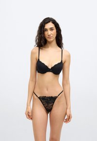 Γυναίκα που φοράει μαύρο δαντελωτό σουτιέν και ταιριαστό δαντελωτό στρινγκ, στέκεται μπροστά σε απλό ανοιχτό φόντο, αντιμετωπίζοντας προς τα εμπρός με χαλαρή στάση.