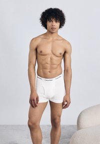 White cotton boxer briefs with a solid design and elastic waistband featuring "Calvin Klein" logo in black. Model has curly hair, standing pose.