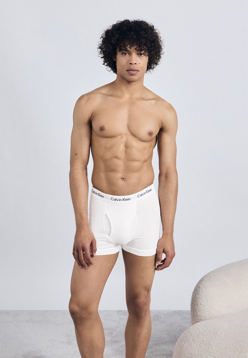 White cotton boxer briefs with a solid design and elastic waistband featuring "Calvin Klein" logo in black. Model has curly hair, standing pose.