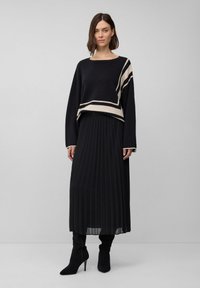 Schwarzer, gekürzter Pullover mit cremefarbenen Streifen, kombiniert mit einem schwarzen, gefalteten Rock. Das Modell trägt schwarze Stiletto-Stiefel und präsentiert einen gestuften Look.
