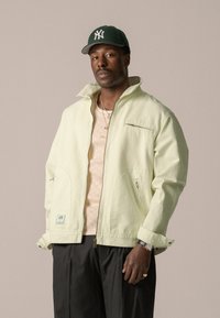 Veste zippée vert clair avec col haut, poches latérales et poche poitrine. Portée sur un haut en tricot beige clair. Pantalon noir.