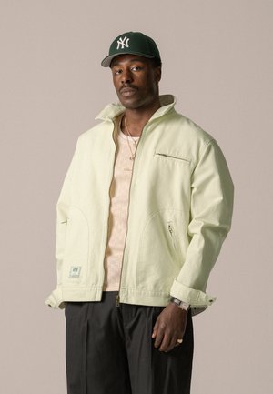 Light green zip-up jacket with high collar, side pockets, and a chest pocket. Worn over a light beige knitted shirt. Black trousers.