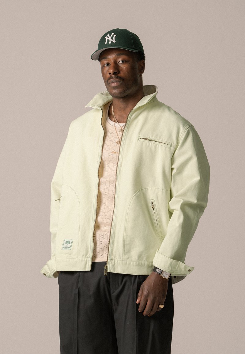 Chaqueta verde claro con cremallera y cuello alto, con bolsillos laterales y un bolsillo en el pecho. Llevada sobre una camiseta de punto beige claro. Pantalones negros.