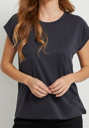 Eine Frau mit welligem braunem Haar hält den Saum eines dunklen T-Shirts und trägt ein einfaches, lässiges Outfit vor einem hellen Hintergrund.