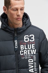 Jonge man met tatoeages die een zwart gewatteerd jack draagt met witte en rode tekst waarop staat "63 Blue Crew David Airboat Tour".