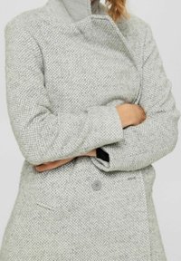 Woman wearing a textured light gray coat with a high collar, buttoned front, and arms crossed across the chest.