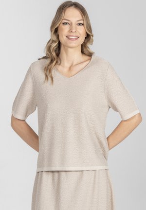 Kobieta o blond, falowanych do ramion włosach, ubrana w beżowy, teksturowany top z krótkim rękawem i dekoltem w serek oraz dopasowaną spódnicę, uśmiechająca się z rękami za plecami.