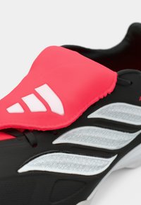 Μαύρο αθλητικό παπούτσι με έντονη ροζ γλώσσα, ασημένιες λεπτομέρειες, υφασμάτινη επιφάνεια και κομψό σχέδιο, διαθέτοντας τρεις λευκές ρίγες στο πλάι.