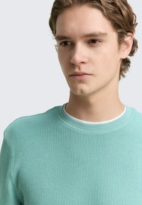 Helle mintgrüne, gerippte Pullover mit rundem Halsausschnitt, über einem weißen Hemd getragen, mit strukturiertem Stoff und dezenten Design-Details.