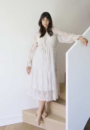 Naine valges pitsist pikkade varrukatega midi-kleidis seisab puidust trepil, toetab kätt valgel piirdel ja kannab beežeid kontsakingi.