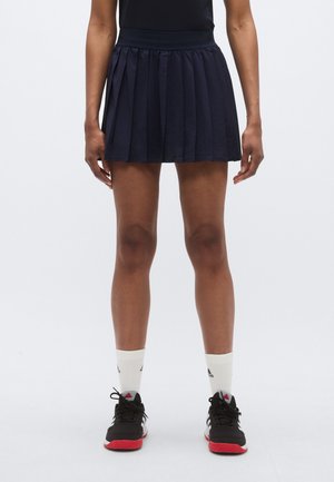 Jupe plissée marine avec une texture douce, taille élastique et longueur mi-cuisse. Associée à des chaussures de sport noires et des chaussettes blanches montantes.