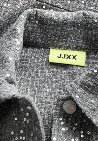 Sivi teksturirani materijal s raspršenim srebrnim šljokicama, s ovratnikom i zelenom etiketom koja glasi "JJXX." Vidljiv detalj gumba.